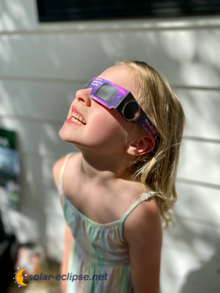 a girl wearing solar eclipse glasses from solar-eclipse.net looking up at the sun